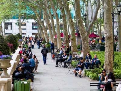 Park users walk down a promenade in Bryant Park in New York City, amid towering trees, a stone balustrade, planters with flowers, and ivy beds.