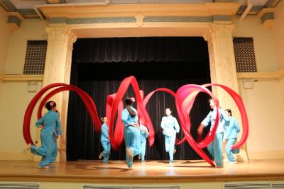 Dancers perform on stage with large red ribbons.