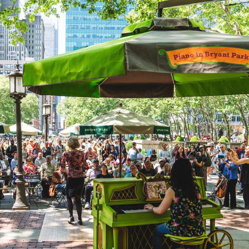 A woman taps while a pianist plays the piano at Bryant Park NYC in front of a large audience outdoors.