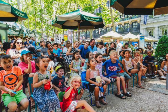 Children seated theatre style in the gravel area of the carousel in Bryant Park looking at awe and shock at the performer on stage